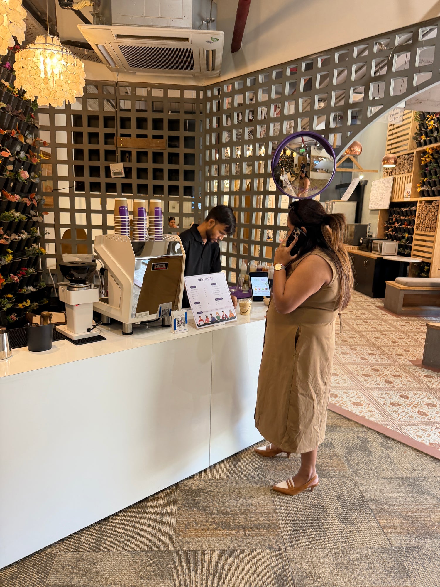 A woman in a beige sleeveless dress stands at a out of office coffee counter inside a cafe, talking on her phone. A barista is visible behind the counter, and the shop has a geometric tiled wall, patterned rug, and decorative lighting.
